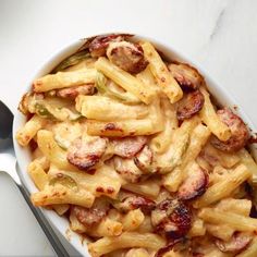 a white bowl filled with pasta covered in sausage and sauce next to a silver spoon