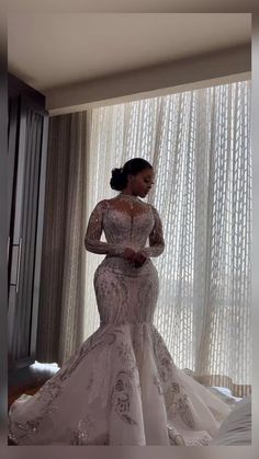 a woman standing in front of a window wearing a wedding dress with long sleeves and beading