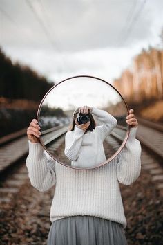 a woman holding a mirror over her face while standing on train tracks