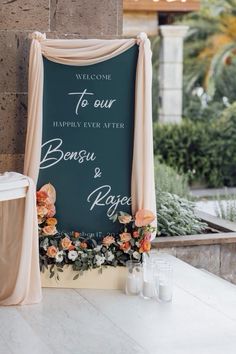 a welcome sign with flowers and greenery on the table at a wedding in front of a stone wall