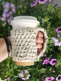 someone is holding a coffee cup in their hand with some flowers behind them and purple flowers