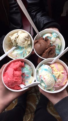 four bowls of ice cream with spoons in them on a table next to two people's hands