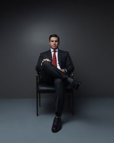 a man in a suit sitting on a chair with his legs crossed and wearing a red tie