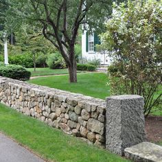 a stone wall in front of a house with trees and grass on the side walk