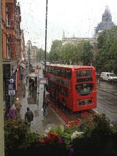 a red double decker bus driving down a street next to tall buildings in the rain