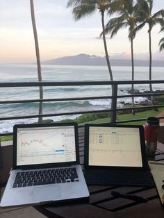 two laptops are sitting on a table near the ocean with palm trees in the background