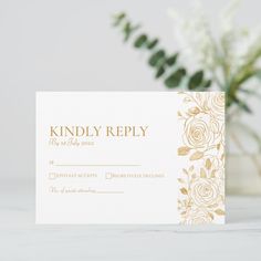 a white and gold wedding response card on a table with some flowers in the background