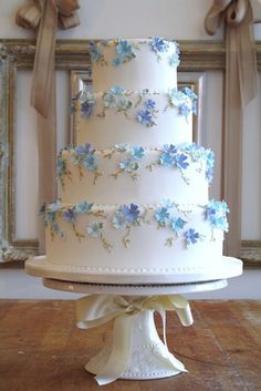 a three tiered white wedding cake with blue flowers on the top and ribbon around the edges