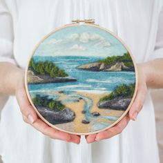 a woman is holding a small hand embroidered landscape with water and land in the background