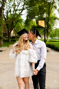 a man and woman in graduation gowns kissing each other on the cheek with trees in the background