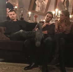 two men and a woman sitting on a couch in a living room with candles behind them