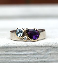 This is a multistone birthstone ring. This particular ring features an amethyst, aquamarine and diamond, but it can be made with other combinations of stones and the shapes could also be changed.If you get in touch we can work out options and prices for you. The ring is recycled sterling silver in a 4mm wide band. The pear shaped amethyst is 5x7mm and the aquamarine is 4mm round with a 1.5mm diamond, all set in recycled 9ct yellow gold. The ring is made to order in the size you need. You can lea Wide Band Mothers Ring, Family Birthstone Ring Ideas, Modern Ring Design Women, Modern Multi-stone Gemstones For Anniversary, Blue Amethyst Anniversary Ring, Modern Multi-stone Birthstone Ring For Anniversary, Modern Topaz Ring With Gemstone Accents For Anniversary, Modern Amethyst Birthstone Ring For Anniversary, Anniversary Multi-stone Amethyst Diamond Ring