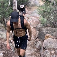 a man with no shirt on walking up a trail in the woods wearing shorts and a hat