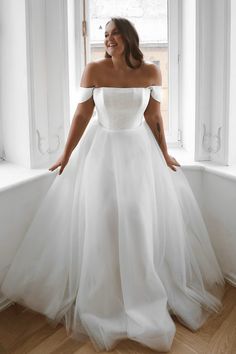 a woman in a white wedding dress standing by a window with her hands on her hips