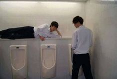 two young men standing next to urinals in a bathroom with one laying on the floor