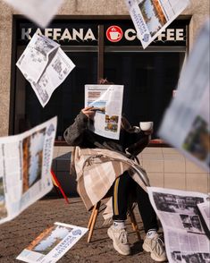 a person sitting on a bench reading a newspaper with papers flying around her and holding a cup