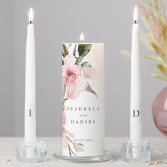 two white candles with pink flowers on them next to a vase and glass candle holder