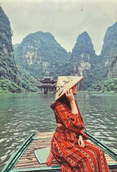 a woman in a red dress and hat sitting on a boat with mountains in the background