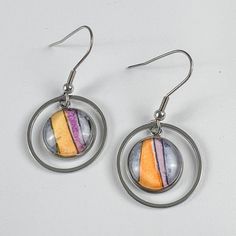 Round watercolor earrings featuring vertical orange, purple and blue stripes. A delicate circular metal frame surrounds the main part of the drop earrings. Multicolor Nickel-free Retro Jewelry, Retro Multicolor Nickel-free Jewelry, Retro Circle Jewelry Gift, Multicolor Round Retro Jewelry, Retro Multicolor Round Jewelry, Artistic Multicolor Hypoallergenic Jewelry, Modern Hand Painted Multicolor Jewelry, Hand Painted Multicolor Circular Jewelry, Bright Watercolor