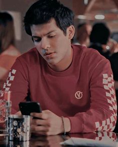 a young man sitting at a table looking at his cell phone while holding a drink