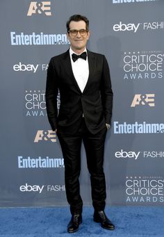 a man in a tuxedo and bow tie standing on a blue carpet with his hands in his pockets