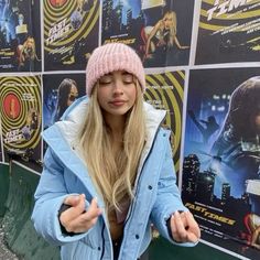 a woman standing in front of a wall with posters on it's sides and wearing a pink beanie