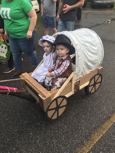 two small children in a wagon on the street with people standing around and looking at them