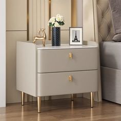 a nightstand with two drawers and a vase on the top, next to a bed