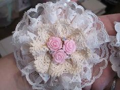 a hand holding a pink and white flower brooch with lace on it's sides