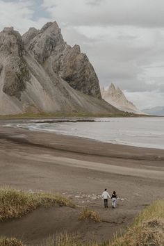 two people walking on the beach with mountains in the backgrouds and water behind them