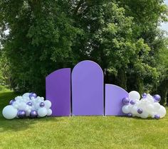 purple and white balloons are in front of an arch on the green grass with trees