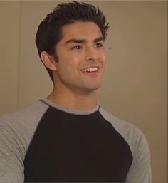 a young man smiles while standing in front of a wall and wearing a gray and black shirt