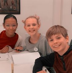 three children are sitting at a table with some pens and paper in front of them