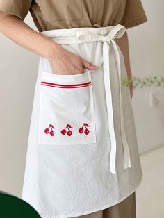 a woman wearing an apron with cherries embroidered on the front and side, while holding her hands in her pocket