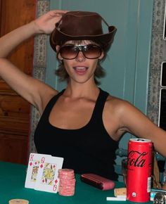 a woman wearing a cowboy hat and sunglasses at a table with poker chips, cards, and a drink