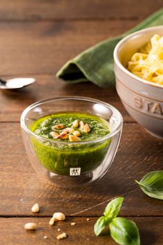 a bowl of pesto next to a bowl of pasta with basil leaves on the side