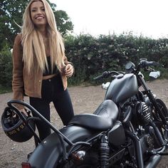 a woman standing next to a black motorcycle on gravel road with trees in the background