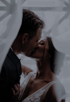 a man and woman kissing in front of a white curtain, with the bride's veil draped over her head