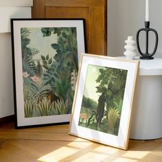 two framed pictures sitting on top of a wooden floor next to a vase and lamp