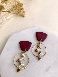 two red and gold earrings sitting on top of a white marble slab next to dried flowers