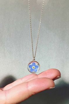 a hand holding a tiny blue flower in a glass ball on a silver chain necklace