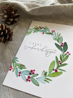 a christmas card with watercolor leaves, berries and pine cones on the table next to it