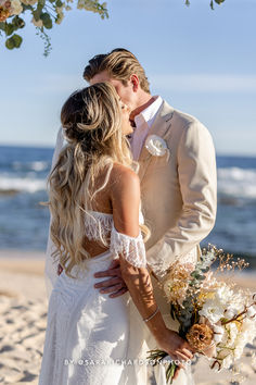 a bride and groom kissing on the beach in front of an arch with flowers hanging from it