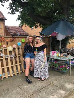 two women standing next to each other in front of a table with an umbrella on it