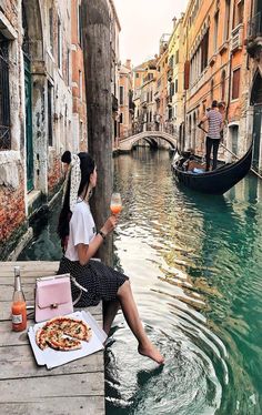 a woman sitting on a dock next to a river with pizza and drinks in front of her