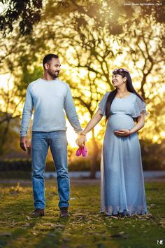 a pregnant couple holding hands and walking through the grass at sunset with trees in the background