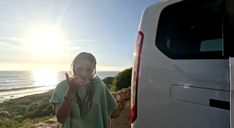 a woman standing next to a van with the sun in the background and water behind her