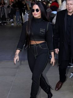 a woman in black outfit walking next to a man wearing sunglasses and a suit with high waist