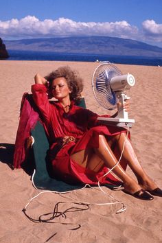 a woman sitting in a chair on the beach with an old fashioned fan next to her