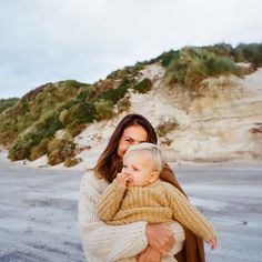 a woman holding a baby in her arms while standing on top of a sandy beach